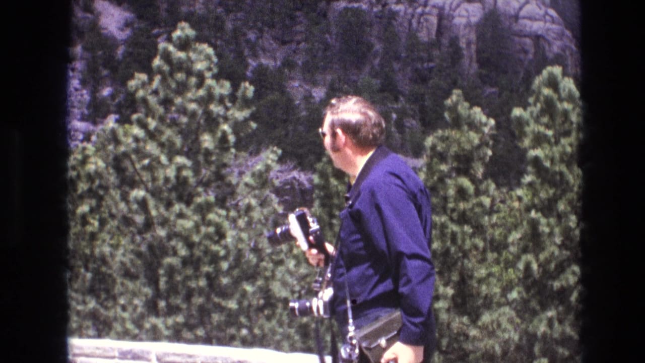 The man turns toward the rocky background while holding camera gear, with evergreen trees and a low stone or concrete edge visible in the foreground.