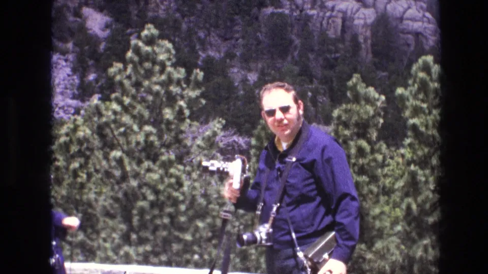The man faces the filming camera in a playful tourist pose, holding a small movie camera while additional camera equipment hangs from his neck and shoulder.