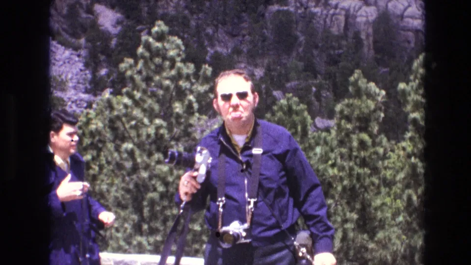 The man mugs toward the camera with his camera gear visible, while another adult man briefly appears in the background against trees and rock.