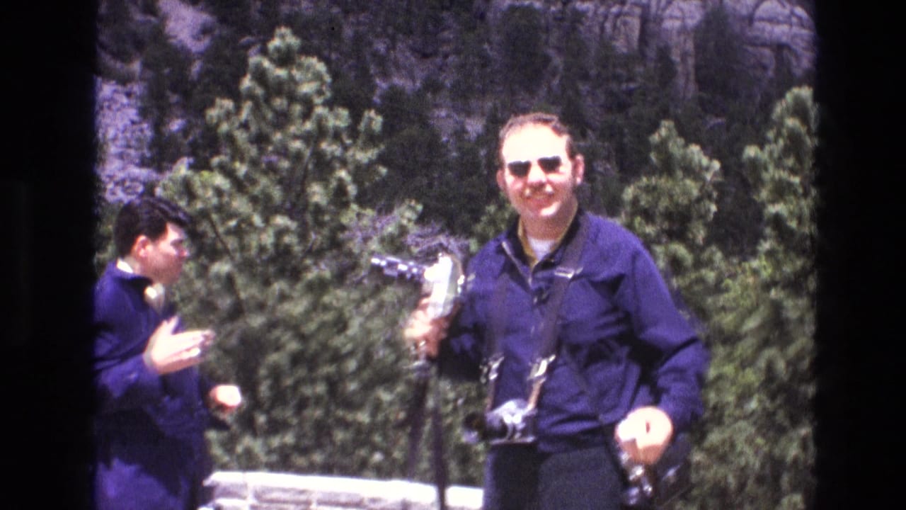 1974 South Dakota home-movie frame showing two adult male tourists at Mount Rushmore, including a smiling man in sunglasses holding a camera near evergreen pines.