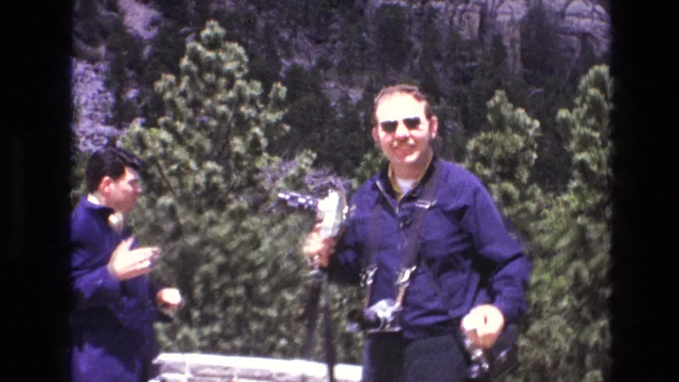 1974 South Dakota home-movie frame showing two adult male tourists at Mount Rushmore, including a smiling man in sunglasses holding a camera near evergreen pines.