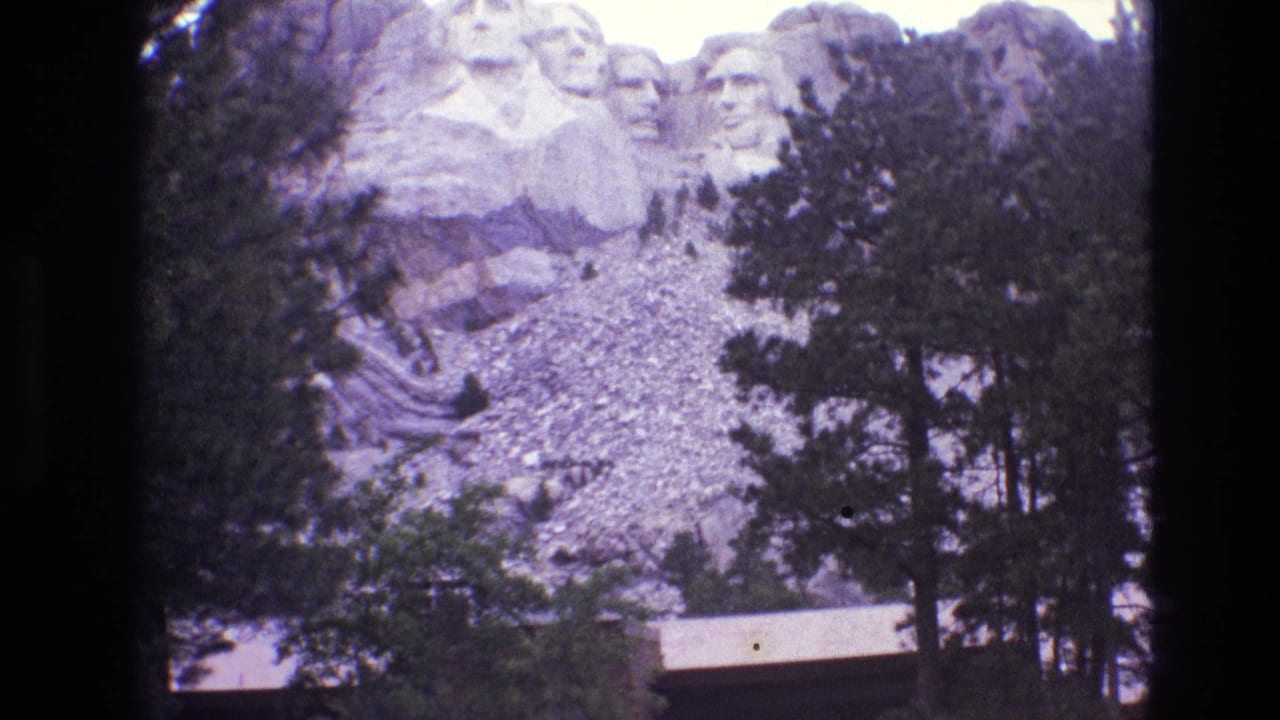 Mount Rushmore viewed through trees, with carved faces, rocky slope, and a low building below.