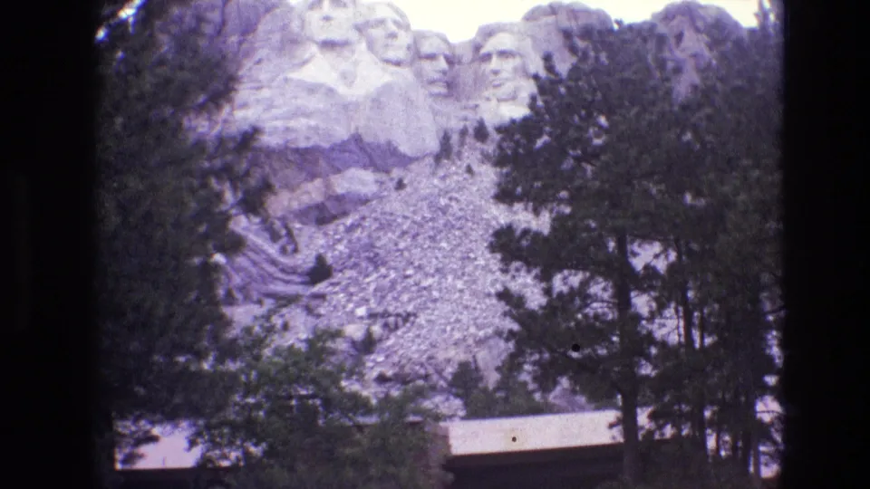 1974 South Dakota home-movie view of Mount Rushmore's carved presidential faces partly framed by evergreen trees and rocky terrain, with a low structure below.