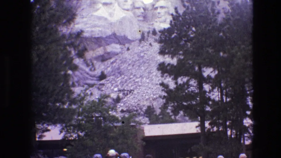 1974 South Dakota home-movie frame of Mount Rushmore behind pines and rocky terrain, with a low structure and small visitor figures near the lower edge.
