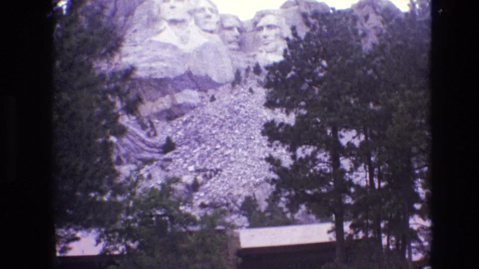 1974 South Dakota 8mm frame showing Mount Rushmore's carved faces beyond evergreen pines, rocky terrain, and a low visitor-area building.