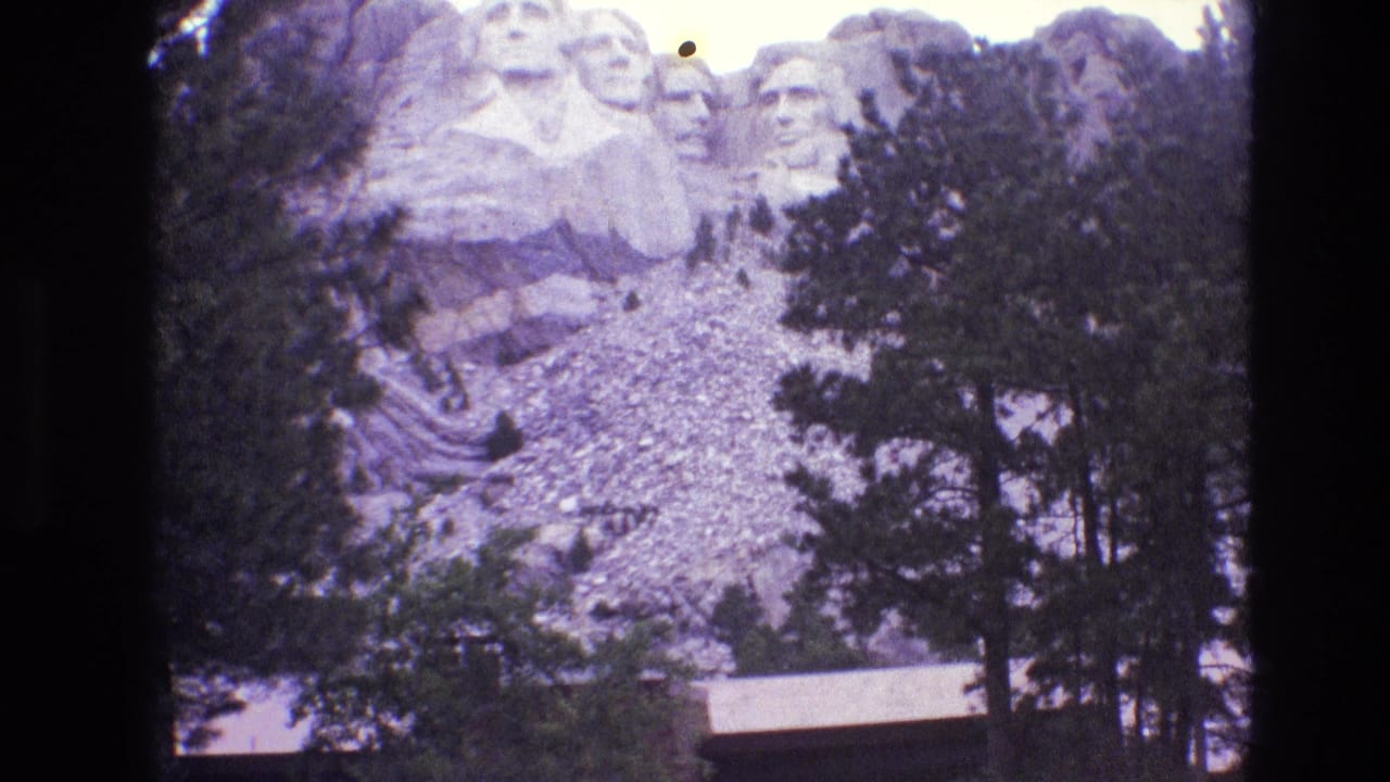 1974 8mm frame of Mount Rushmore in South Dakota, with the presidential carvings partly framed by evergreen trees and a pale rocky slope below.