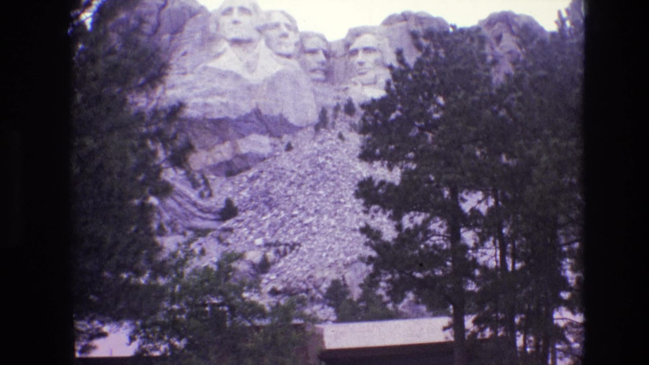 1974 visitor-shot 8mm view of Mount Rushmore, with the carved presidential faces above a tree-lined rocky slope and a small roofline near the bottom edge.