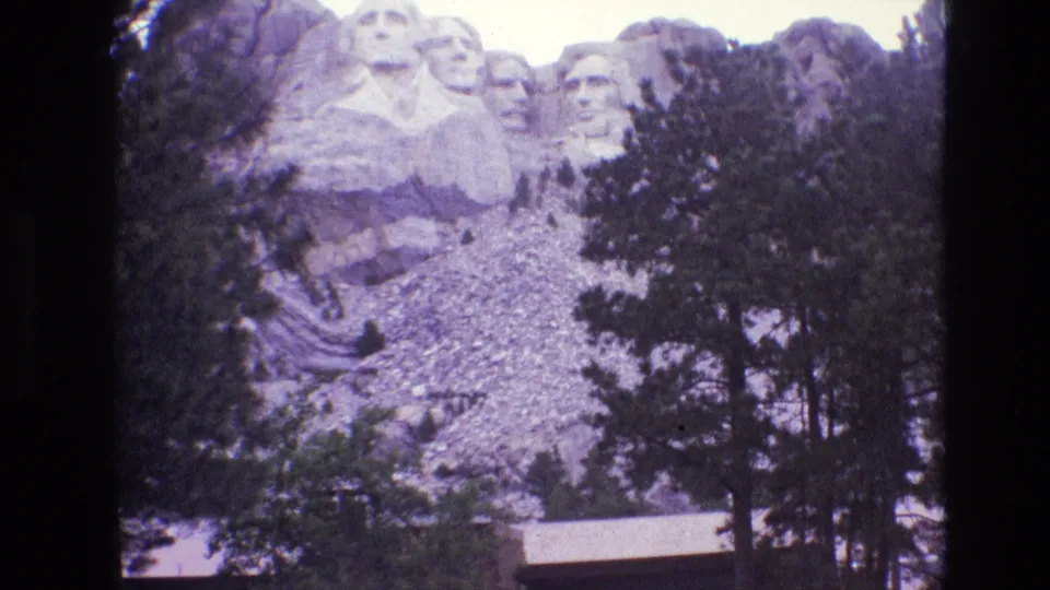1974 visitor-shot 8mm view of Mount Rushmore, with the carved presidential faces above a tree-lined rocky slope and a small roofline near the bottom edge.