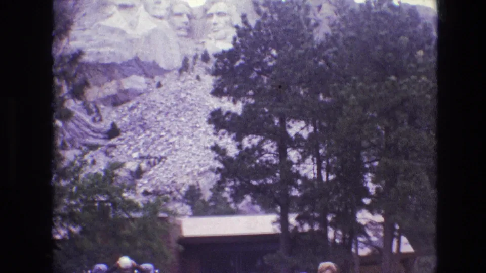 1974 8mm frame looking toward Mount Rushmore from the visitor area, with trees, a low building, and several tourists visible along the lower edge.