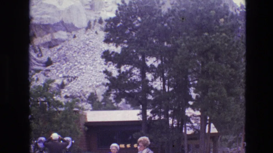 1974 8mm frame of the Mount Rushmore visitor area, with tourists in the foreground, evergreen trees, a low structure, and the rocky monument slope behind them.
