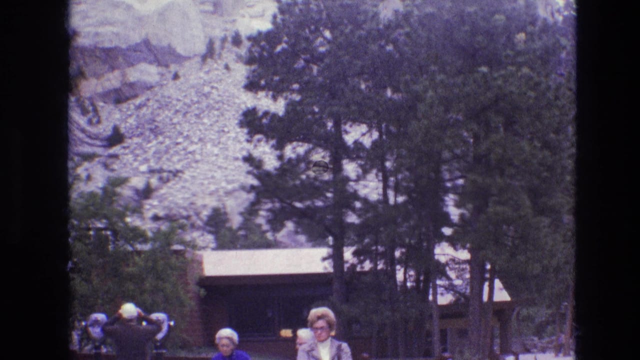 1974 South Dakota 8mm frame showing visitors near tall pines, a rustic wooden structure, and pale rock formations in the background.