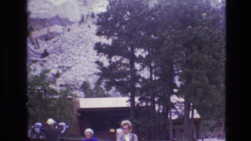 1974 South Dakota 8mm frame showing visitors near tall pines, a rustic wooden structure, and pale rock formations in the background.