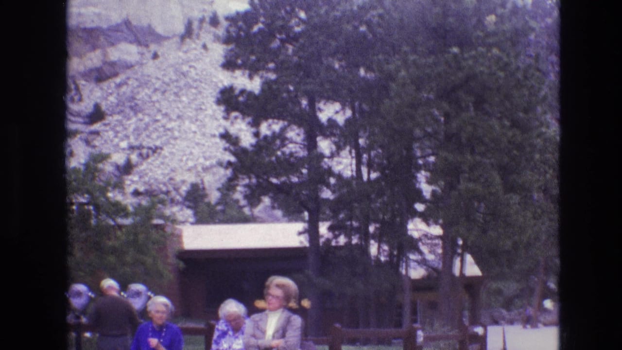1974 South Dakota park visitors stand and look around near a wooden building, with tall pines and pale rock formations behind them.