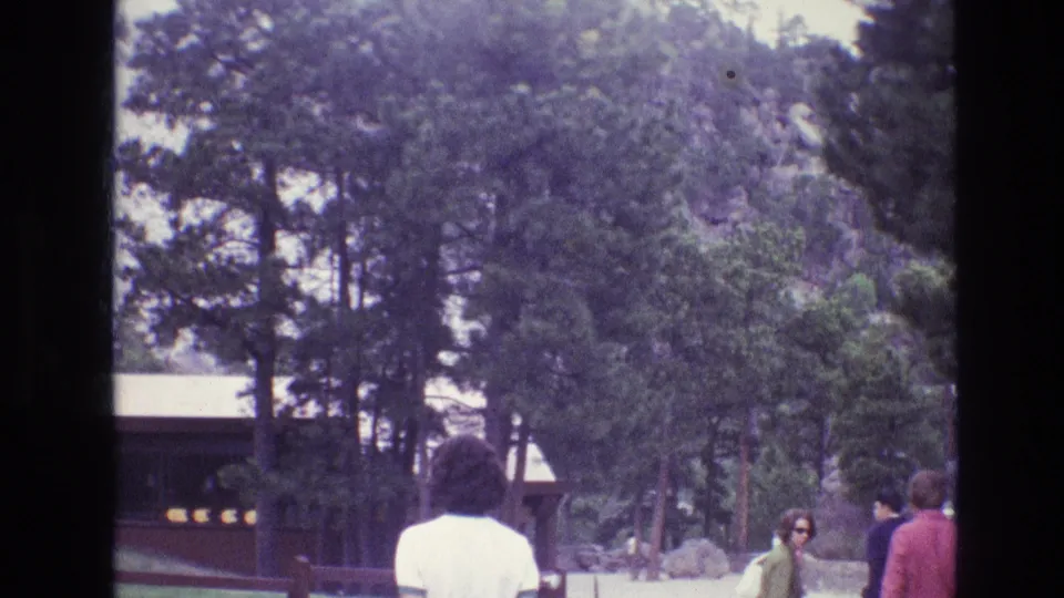 1974 South Dakota home-movie frame showing a visitor walking by a fence line below tall pines and a rustic building.
