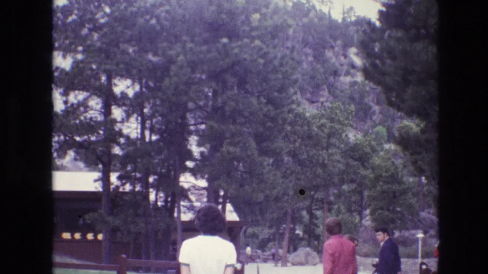 1974 South Dakota 8mm frame with tourists walking through a pine-covered park area beside a fence, rustic building, and rocky slope.