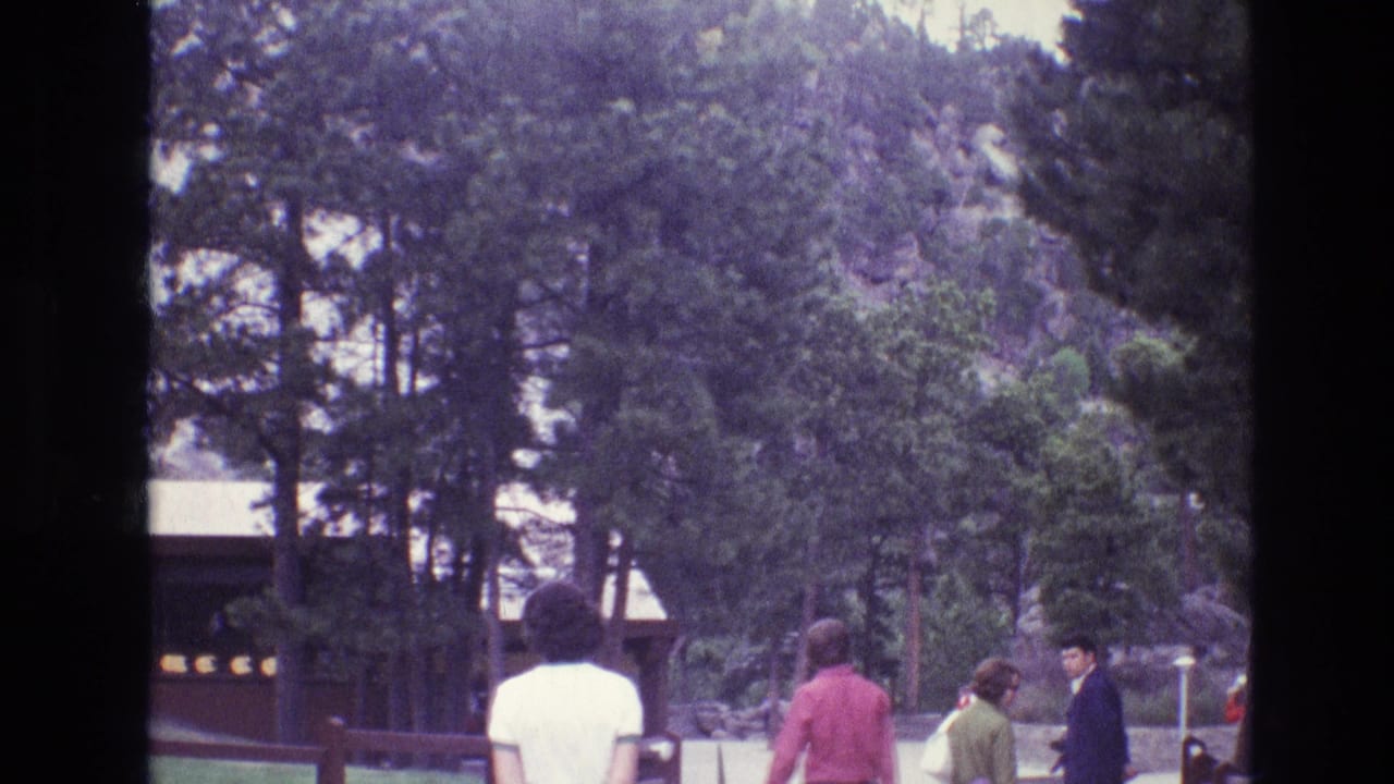 1974 South Dakota 8mm frame showing visitors in period clothing walking through a pine-lined visitor area near a rustic park building at Mount Rushmore.