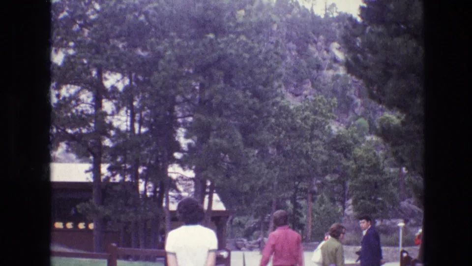 1974 South Dakota 8mm frame showing visitors in period clothing walking through a pine-lined visitor area near a rustic park building at Mount Rushmore.
