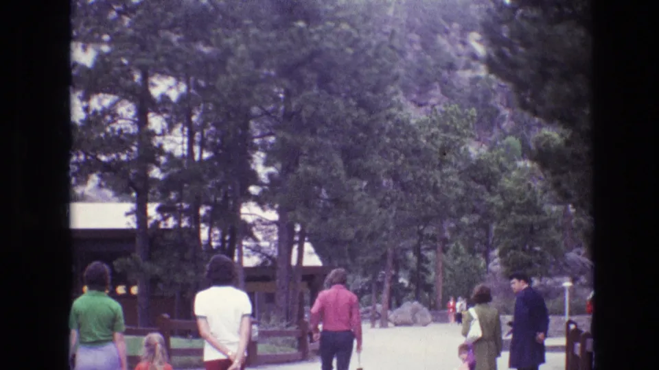 A 1974 home-movie frame shows several visitors walking along the Mount Rushmore grounds, with tall pines, a rustic structure, and park landscaping around them.