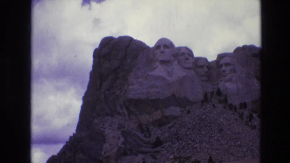 1974 8mm home-movie frame of Mount Rushmore in South Dakota, showing the carved presidential heads in shadow beneath a cloudy sky.