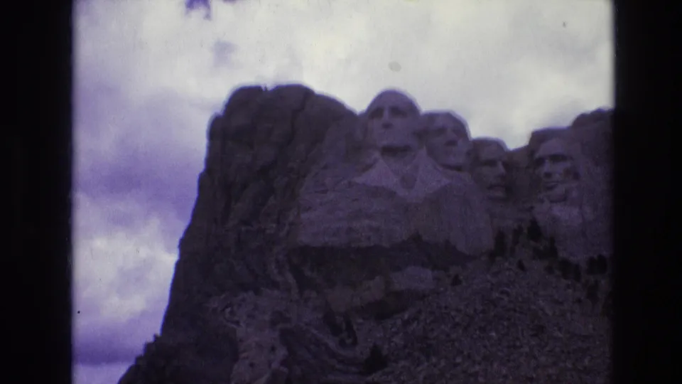 1974 8mm frame showing a slightly soft, purple-tinted tourist view of Mount Rushmore's granite carvings beneath layered clouds.