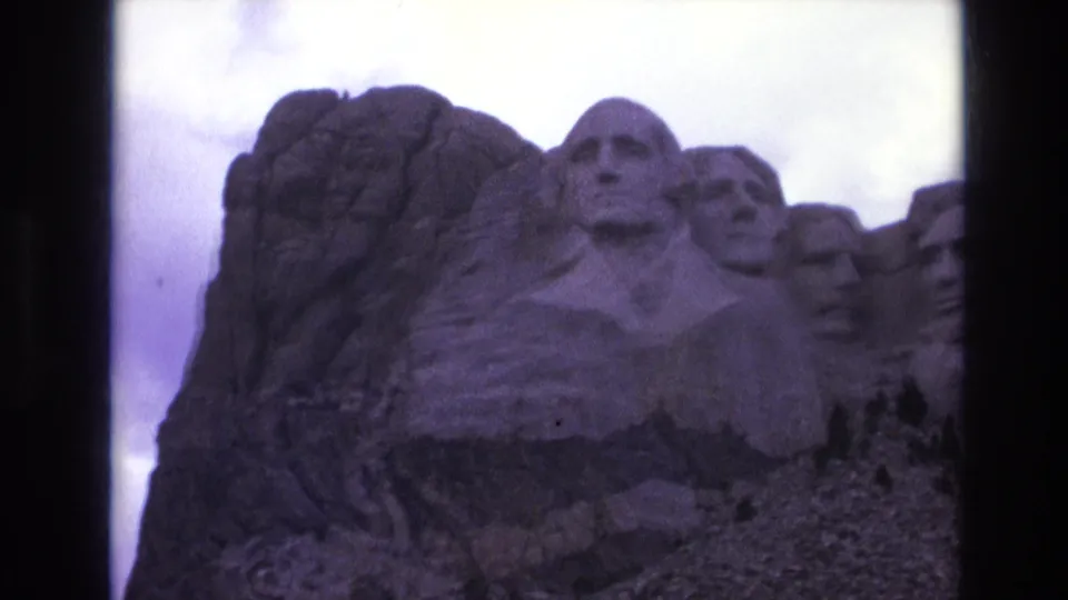 1974 South Dakota home-movie frame with a closer low-angle view of Mount Rushmore, emphasizing Washington's carving and adjacent presidential heads in shadow.