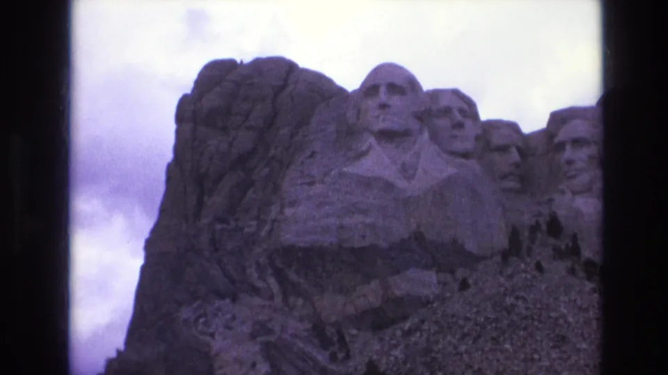 1974 8mm frame of the Mount Rushmore presidential carvings, with the granite cliff and overcast sky visible around the monument.