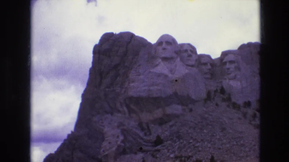 Soft 1974 8mm home-movie view of Mount Rushmore, showing the carved heads above a rocky slope and sparse trees.