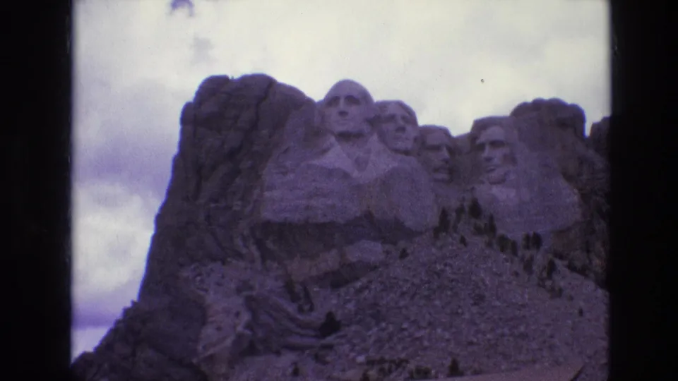 1974 South Dakota home-movie frame showing Mount Rushmore's presidential sculptures, rugged granite, trees, and cloudy sky.