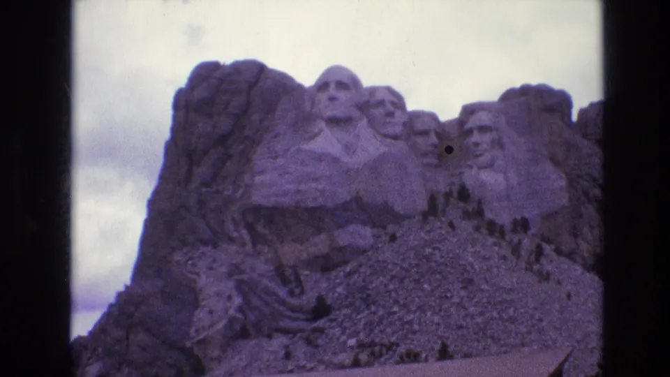 1974 8mm home-movie frame of Mount Rushmore in South Dakota, showing the carved presidential faces with cloudy sky and dark reel borders.