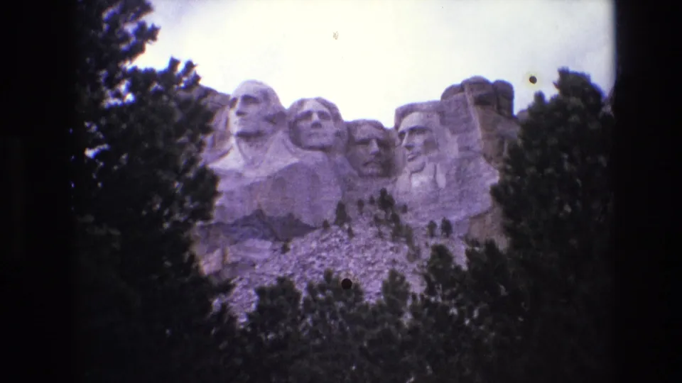 1974 South Dakota 8mm frame showing Mount Rushmore behind dark pine trees, with the carved presidents partly visible against a bright cloudy sky.
