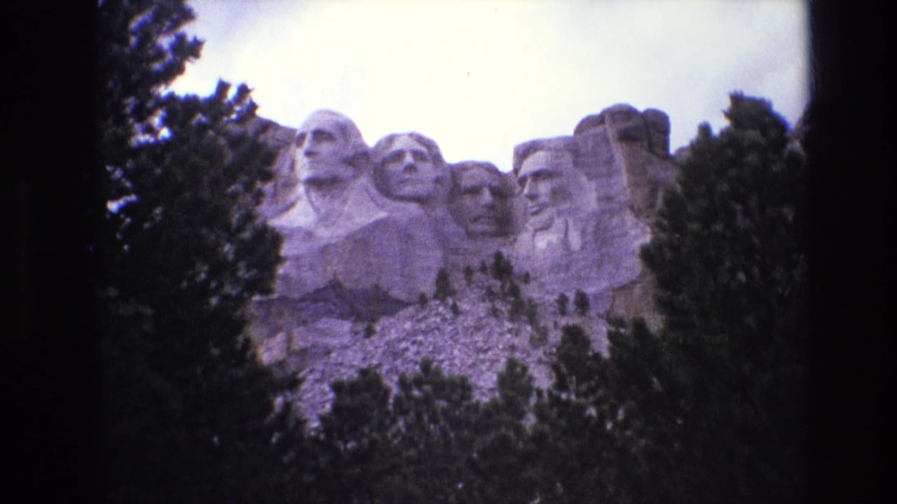 1974 8mm home-movie frame of Mount Rushmore in South Dakota, with the carved presidential heads framed by evergreens and a bright sky.
