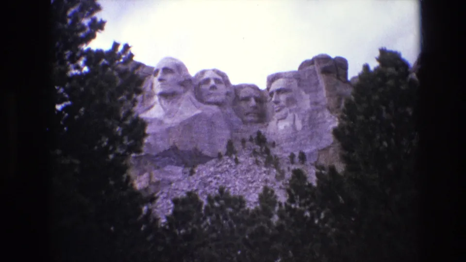 1974 8mm home-movie frame of Mount Rushmore in South Dakota, with the carved presidential heads framed by evergreens and a bright sky.