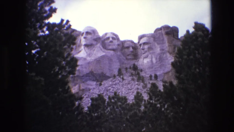 1974 South Dakota visitor footage frame showing the Mount Rushmore carvings partially enclosed by evergreen branches and dark scan edges.