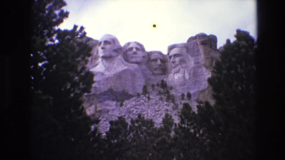 1974 8mm frame showing Mount Rushmore from a tourist viewpoint, with evergreens crossing the lower and side edges of the monument view.