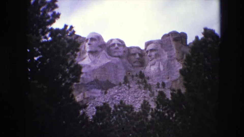 1974 South Dakota home-movie frame with Mount Rushmore centered behind dark evergreen trees, showing soft focus and visible film grain.
