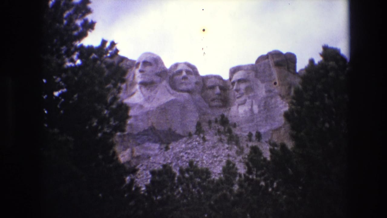 1974 8mm home-movie frame showing a wide visitor view of Mount Rushmore National Memorial in South Dakota, with carved presidential heads partly framed by evergreens.