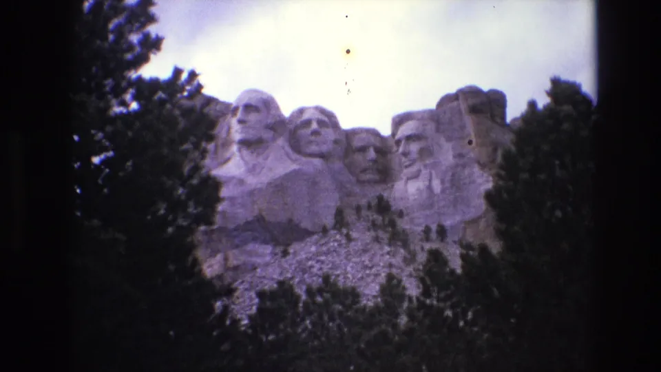 1974 8mm home-movie frame showing a wide visitor view of Mount Rushmore National Memorial in South Dakota, with carved presidential heads partly framed by evergreens.
