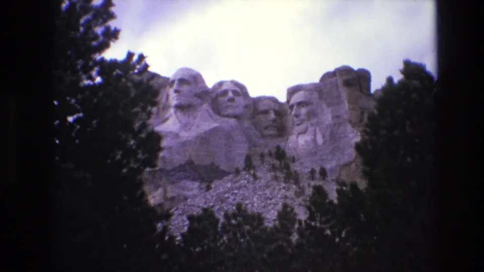 1974 8mm frame of Mount Rushmore seen from a distance, with the presidential carvings centered between evergreen silhouettes and a bright overcast sky.