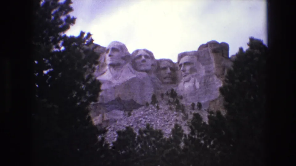 1974 South Dakota home-movie frame showing Mount Rushmore from a lower visitor angle, with trees and broken rock below the carved heads.