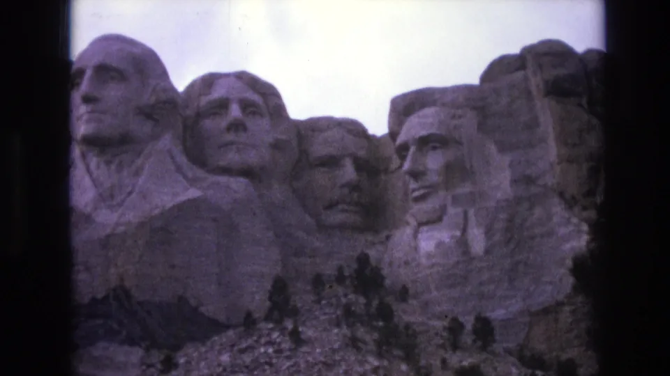Closer 1974 8mm view of Mount Rushmore National Memorial, showing the carved presidential faces above a line of dark trees and rocky foreground.