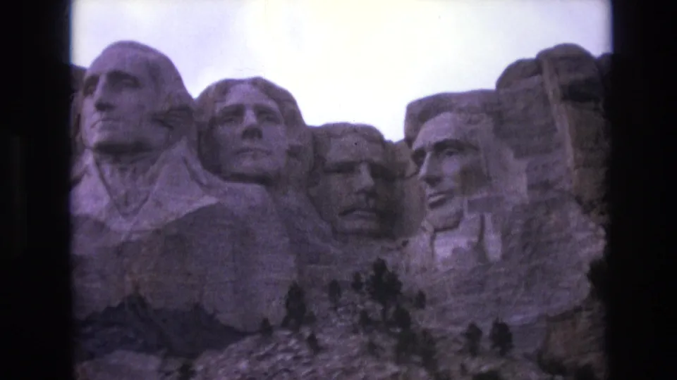 1974 South Dakota 8mm frame showing Mount Rushmore's carved presidential heads under a bright sky, with rocky foreground trees and visible film-frame edges.