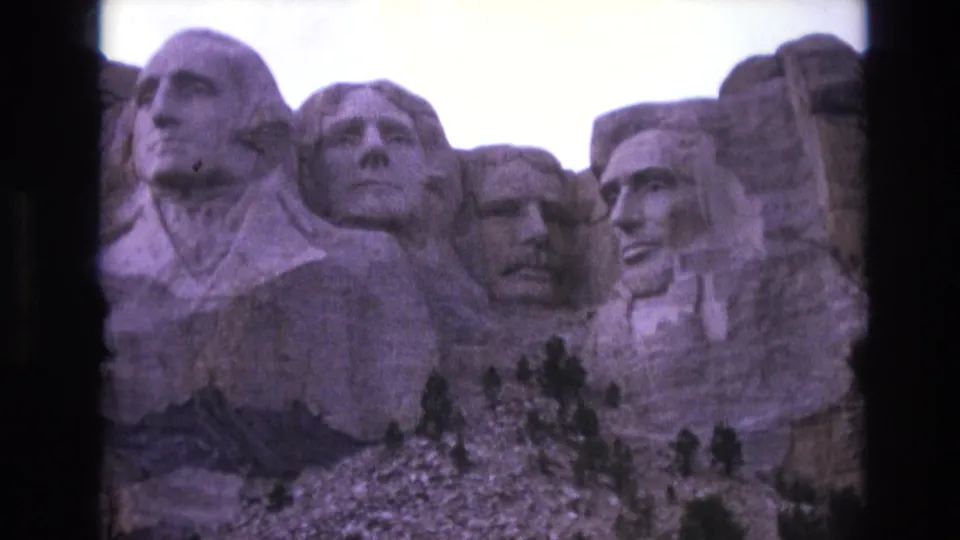 1974 South Dakota tourist-shot 8mm frame with Mount Rushmore's carved heads filling most of the view, with trees and rock below the monument.