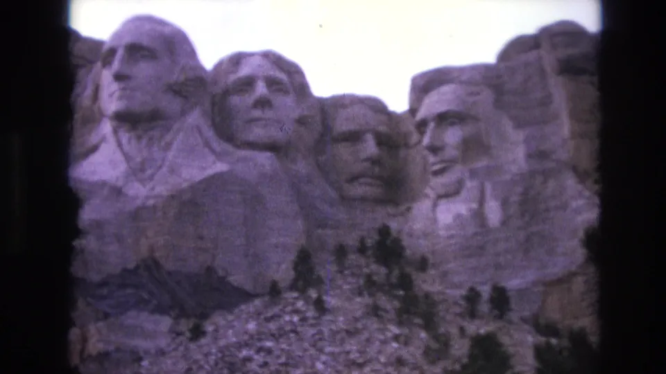 1974 8mm frame showing Mount Rushmore in South Dakota from below, with the presidential carvings, bright sky, rocky base, and small trees in view.