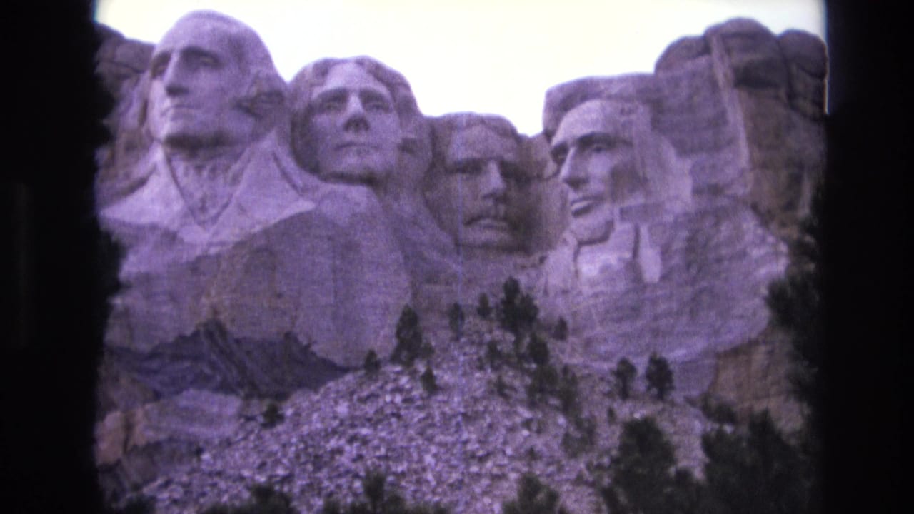1974 8mm home-movie frame showing Mount Rushmore's carved presidential faces from below, with granite slope and trees at the base.