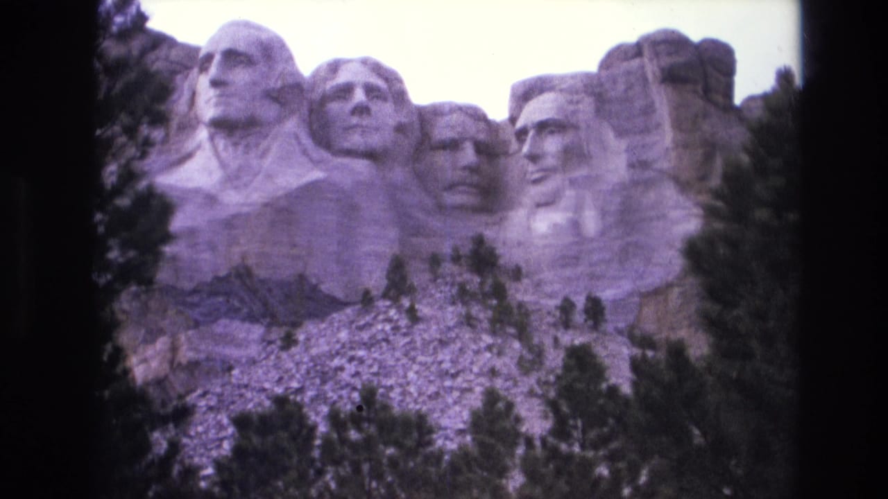 1974 South Dakota home-movie frame with the four carved faces at Mount Rushmore, seen through a soft purple 8mm color cast.