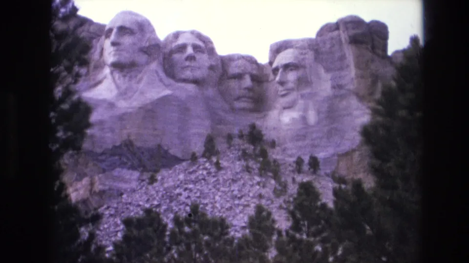 1974 South Dakota home-movie frame with the four carved faces at Mount Rushmore, seen through a soft purple 8mm color cast.