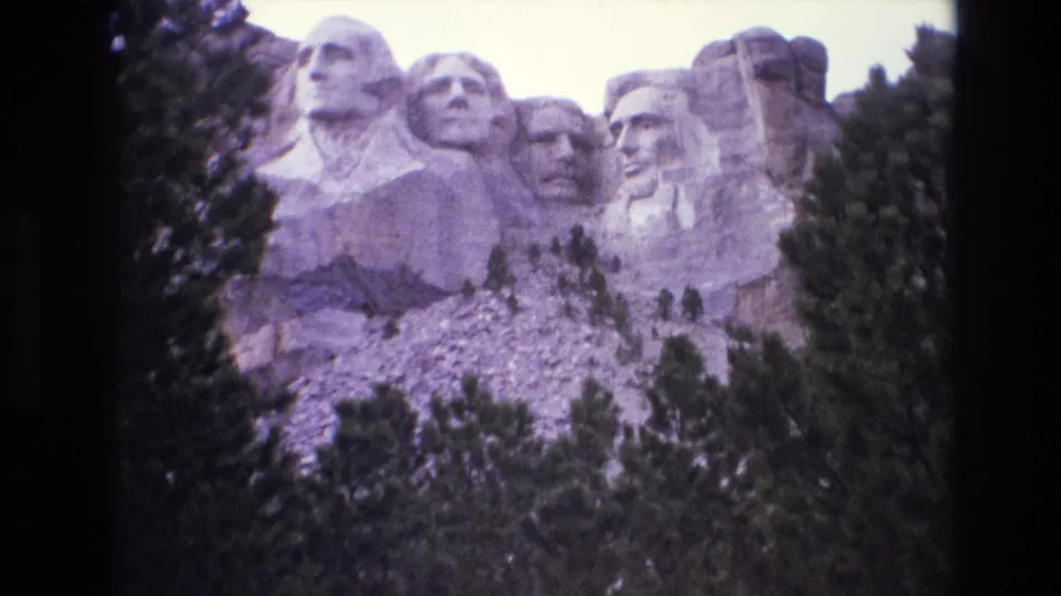 1974 8mm frame of Mount Rushmore from a visitor viewpoint, with dark trees along both sides and the granite monument rising above.