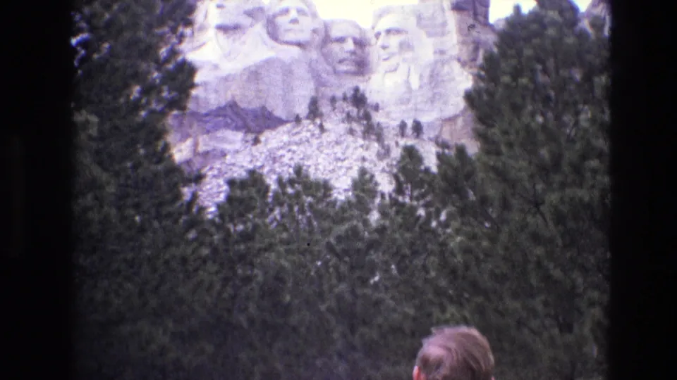 1974 South Dakota 8mm frame showing Mount Rushmore above pine trees, with the back of a visitor's head entering the foreground for scale.