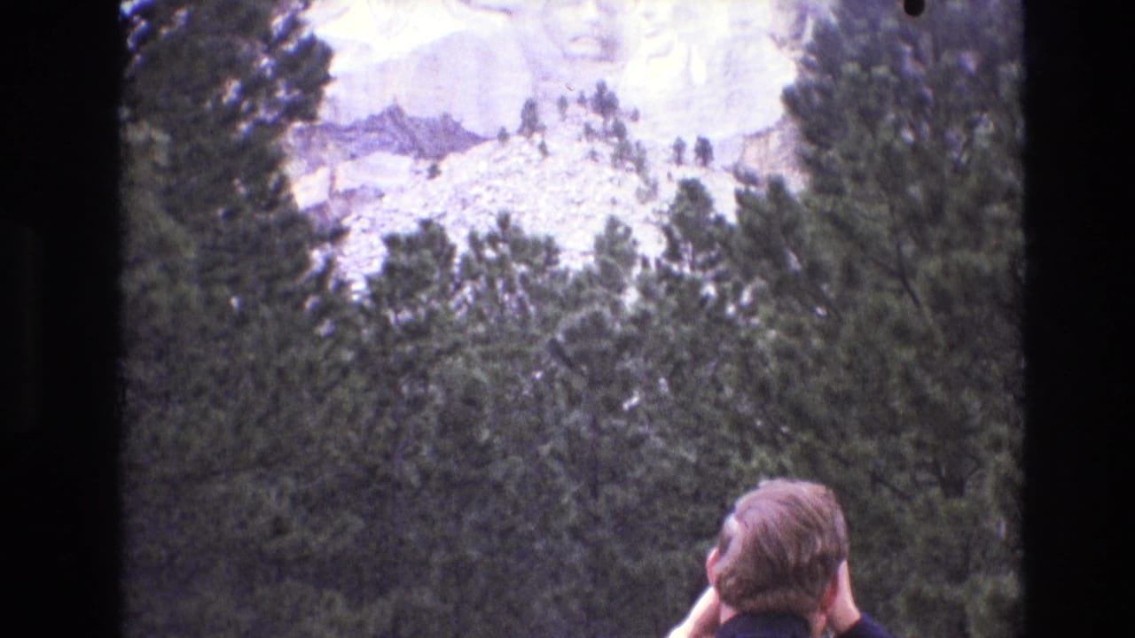 1974 South Dakota home-movie frame showing Mount Rushmore partly framed by pine trees as a man looks upward from the foreground.