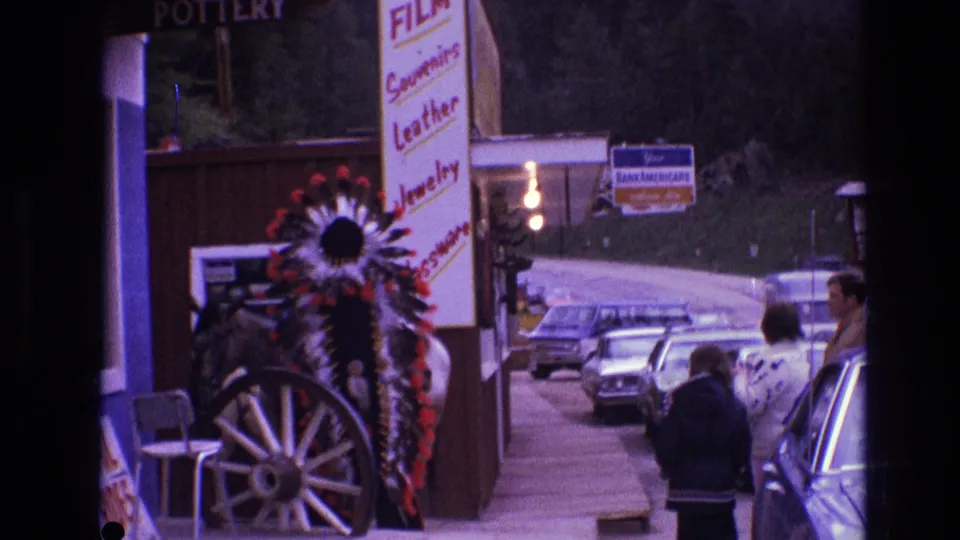 1974 South Dakota street scene with parked cars, a souvenir storefront, visible shop signage, and a Native American headdress display near the sidewalk.